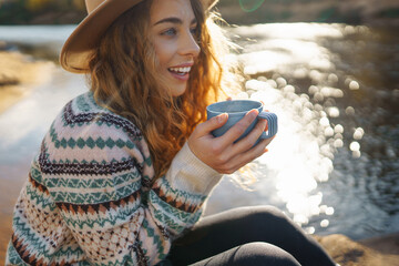 Beautiful woman traveler in a hat sits on a log near the river, drinks a hot drink from a thermos....