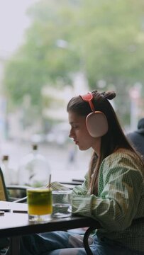 A caf&eacute; backdrop showcases a young woman using headphones and talking on her laptop.