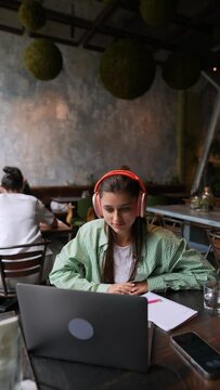 A café setting finds a young lady using headphones while having a conversation on her laptop.