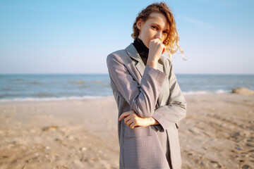 A beautiful woman in stylish clothes stands on the beach on an autumn morning. Fashion model posing among the lush grass on the beach. Fashion and style concept.