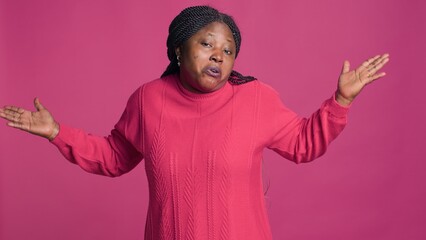 Female fashion blogger standing and looking at camera with baffled expression. Youthful black woman with her hands out motioning no idea no clue illustration against isolated pink background.