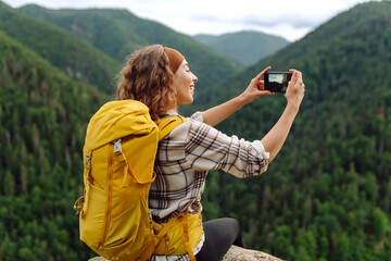 Young traveler with a yellow backpack is resting and taking pictures on the phone on a mountain...