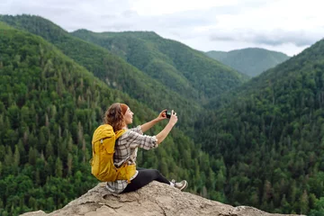 Photo sur Plexiglas Tatras Young traveler with a yellow backpack is resting and taking pictures on the phone on a mountain cliff, admiring the landscape of the high Tatras, mountain range. Beautiful woman on top of the mountain
