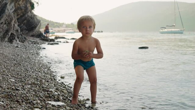 Cute little boy having fun on the sea beach and throwing stones in sea waves.