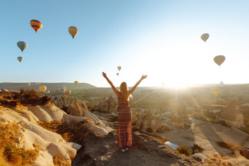 Woman tourist in a hat stands with her back on a mountain, looking at the balloons in Cappadocia,...