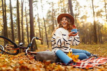 Beautiful curly woman in a hat sits on a red mat with a thermos in the autumn forest. Female enjoys...