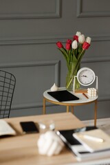 Coffee table with vase of tulips, desk clock, tablet, pen and decorations on desktop background with notebooks and phone in gray room