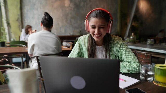 A young woman with headphones is talking on her laptop in a café.