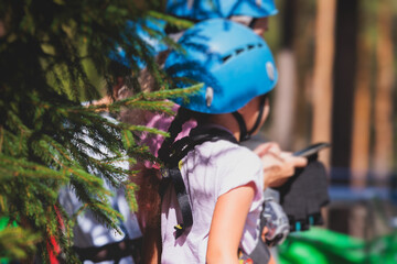 View of high ropes course, kids of climbing in amusement acitivity rope park, passing obstacles and zip line on heights, children teenagers in equipment gear between the trees on heights, summer day