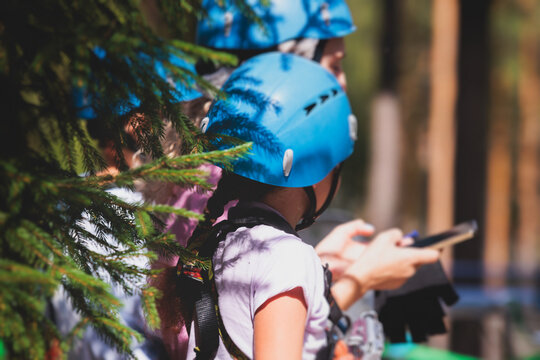 View of high ropes course, kids of climbing in amusement acitivity rope park, passing obstacles and zip line on heights, children teenagers in equipment gear between the trees on heights, summer day
