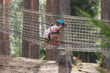 View of high ropes course, kids of climbing in amusement acitivity rope park, passing obstacles and zip line on heights, children teenagers in equipment gear between the trees on heights, summer day