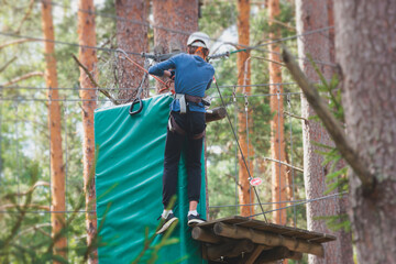 View of high ropes course, kids of climbing in amusement acitivity rope park, passing obstacles and zip line on heights, children teenagers in equipment gear between the trees on heights, summer day