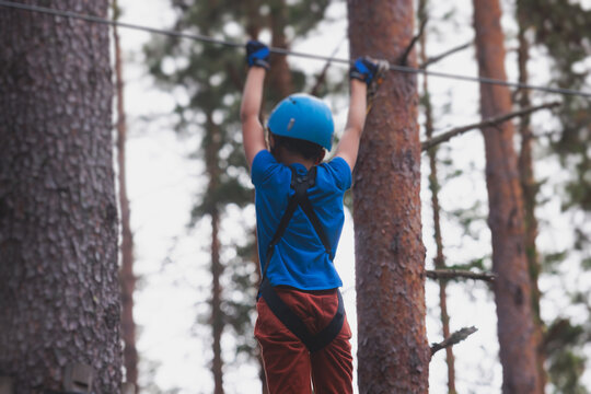 View of high ropes course, kids of climbing in amusement acitivity rope park, passing obstacles and zip line on heights, children teenagers in equipment gear between the trees on heights, summer day