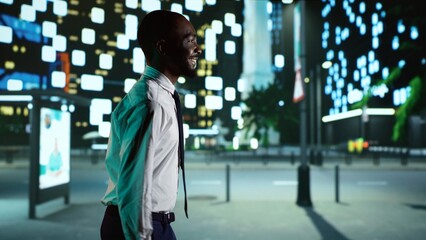 African american man walks downtown at night and admiring urban modern skyscrapers under city...