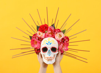 Female hands holding painted human skull for Mexico's Day of the Dead (El Dia de Muertos) with flowers on yellow background