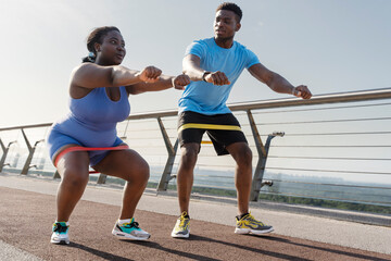 African american body positive woman in sportswear exercising outdoors, squatting with fitness band