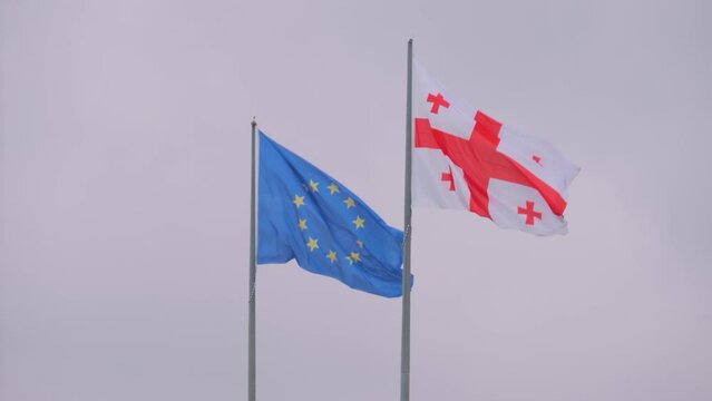 Colorful Georgian and European Union flags are fluttering in the wind against gray sky in Georgia, overcast - slow motion. Diplomacy, politics, patriotism, freedom and international affairs concept