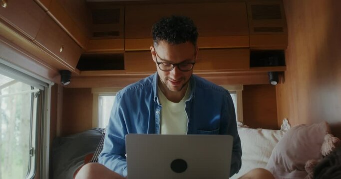 A young man with glasses uses a laptop sitting on a bed, traveling on a RV
