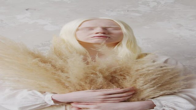 Vertical zoom in studio portrait of young albino woman standing with eyes closed holding dried bouquetVertical zoom in studio portrait of young albino woman standing with eyes closed holding dried bou