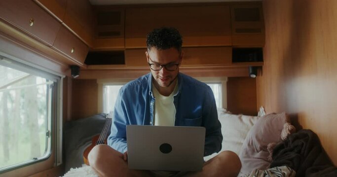 A young man with glasses uses a laptop sitting on a bed, traveling on a RV