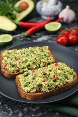 Slices of bread with tasty guacamole and ingredients on black textured table, closeup