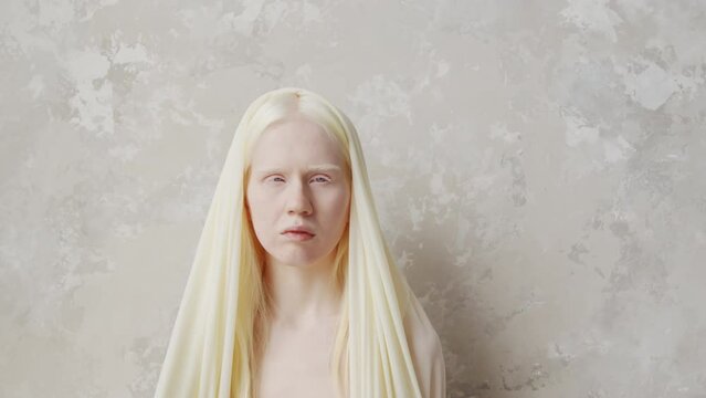 Contemporary studio portrait of shirtless young albino woman wearing veil standing against white wall background