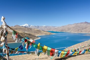 Behold the serene splendor of Lake Yamdrok Tso in Tibet, framed by colorful Tibetan prayer flags set against the enchanting azure hue of the lake's waters.