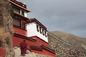 Fototapeta na wymiar Set against a backdrop of towering, snowless mountains, a solitary Tibetan monk clad in traditional robes ascends a rugged path towards the ancient Drigung Thil Monastery.