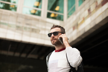 A young, traveling man wearing sunglasses, talking on a smartphone phone, on the street, in a city in Spain.
