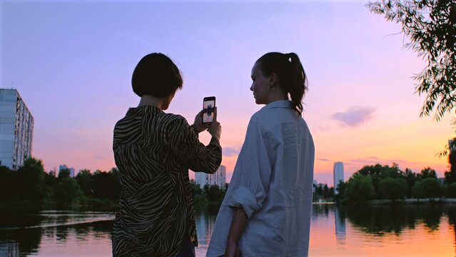 Two women take photos of lake on camera. Beautiful girls taking video of sunset on mobile phone. Female couple friends in summer evening, nature landscape