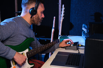 Musician man adjusting the sound of his electric guitar with equalizer, headphones and laptop at home recording studio. Music production.