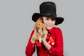 A magician boy in a black hat is holding a red rabbit.