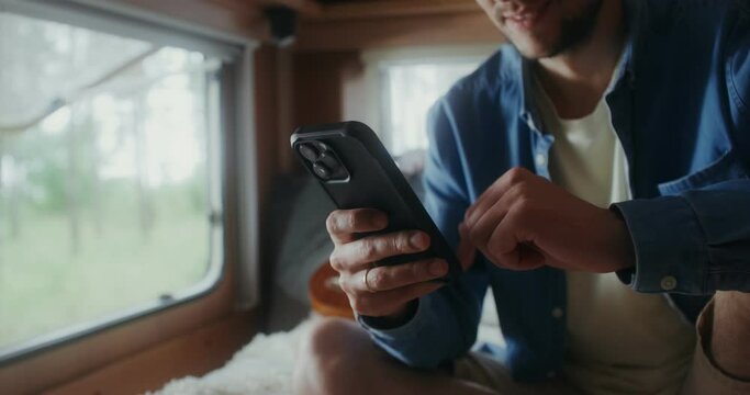 A young man with glasses smiles while using a mobile phone while sitting on a bed in a van. Close-up of his hands
