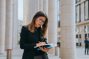 Young adult businesswoman in black jacket writing in notebook stands outside at huge building,...