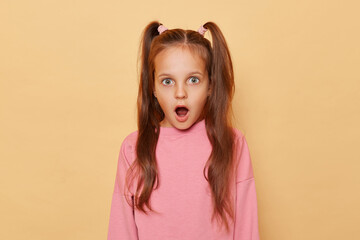 Shocked surprised little girl with ponytails wearing pink sweatshirt isolated over beige background...