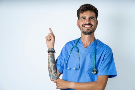 Smiling young caucasian doctor man wearing blue medical uniform indicating finger empty space showing best low prices