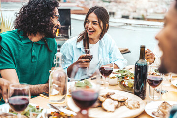 Multiethnic friends having fun at rooftop bbq dinner party - Group of young people diner together sitting at restaurant dining table - Cheerful multiracial teens eating food and drinking wine outside