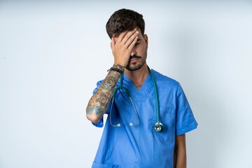 Tired overworked young caucasian doctor man wearing blue medical uniform has sleepy expression, gloomy look, covers face with hand, has eyes shut, gasps from tiredness, fatigue after party