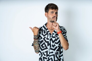 young caucasian man wearing printed shirt asking to be quiet with finger on lips pointing with hand to the side. Silence and secret concept.