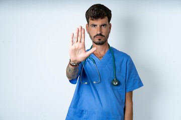 young caucasian doctor man wearing blue medical uniform shows stop sign prohibition symbol keeps...