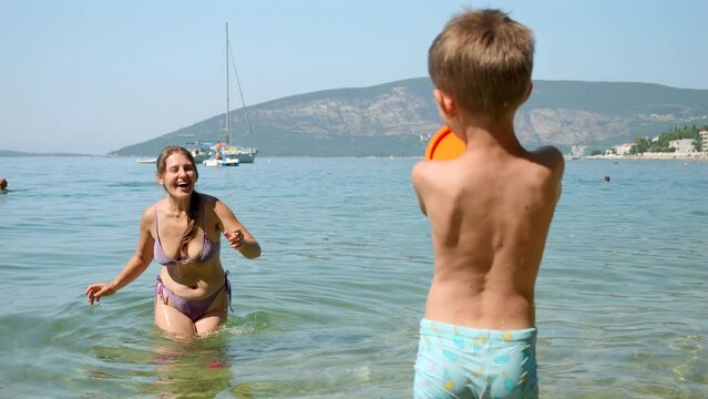 Slow motion of little boy throwing frisbee to his mother standing in the sea next to the beach. Family holiday, vacation and fun summertime of children and parents