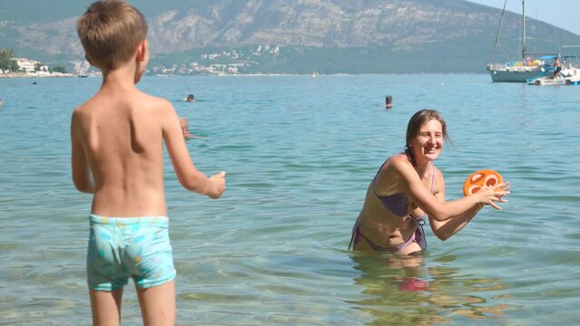 Slow motion of cheerful boy with mother throwing and catching frisbee disk while relaxing in the calm sea water at beach. Family holiday, vacation and fun summertime of children and parents