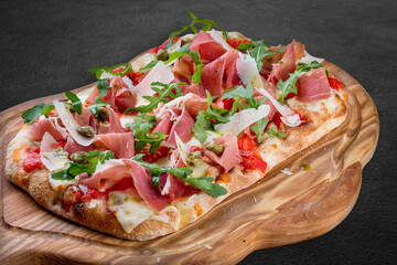 Sorrentina pizza with prosciutto, arugula, capers, pelati sauce, pesto. Roman pizza rectangular on dark background