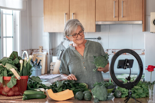 Senior Attractive Woman Streaming Online By Smartphone Follows A Vegetarian Cooking Class. Home Kitchen Table Full Of Vegetables, Healthy Eating Concept