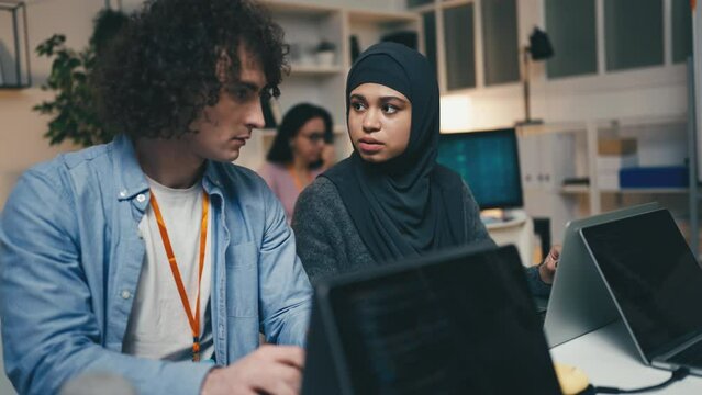 Female programmer in hijab talking with a male colleague in office, diverse team