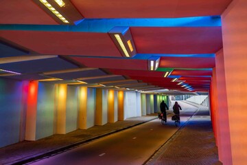 Two cyclists ride side by side through a colorfully lit bicycle tunnel