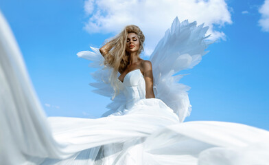Fototapeta na wymiar Beautiful sexy blonde woman in white long elegant dress and angel wings is sensually posing against the heaven on summer day. Angel woman.