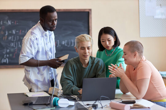 Group of young intercultural students or IT engineers discussing online data or decoding it while sitting in front of laptop in classroom