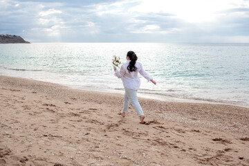 beautiful plus size woman runinf  in the beach against the sea. The fat woman holding the bouquete of whitr flowers. The overweight model wearing style shirt and jeans