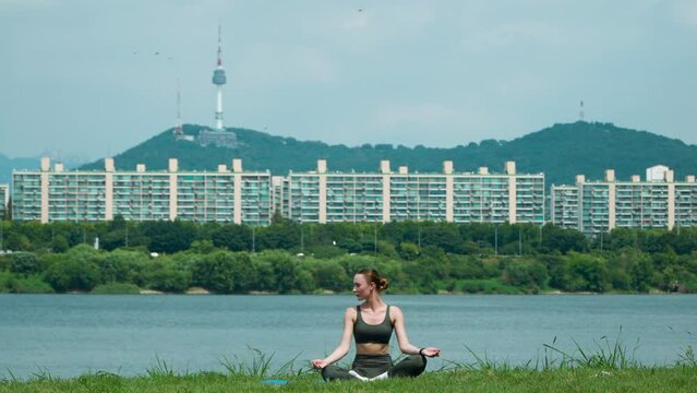 Slender young woman sitting on yoga mat in lotus position and raising hands up outside in Han river city park. Front view of female practicing yoga outdoors on sunny day, N Seoul Namsan Tower View